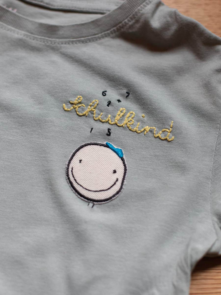 Ein Shirt in graublau liegt auf dem Tisch und darauf ist ein Smiley Aufnäher von halfbird mit Beinen und darüber ist das Wort Schulkind gestickt und zahlen