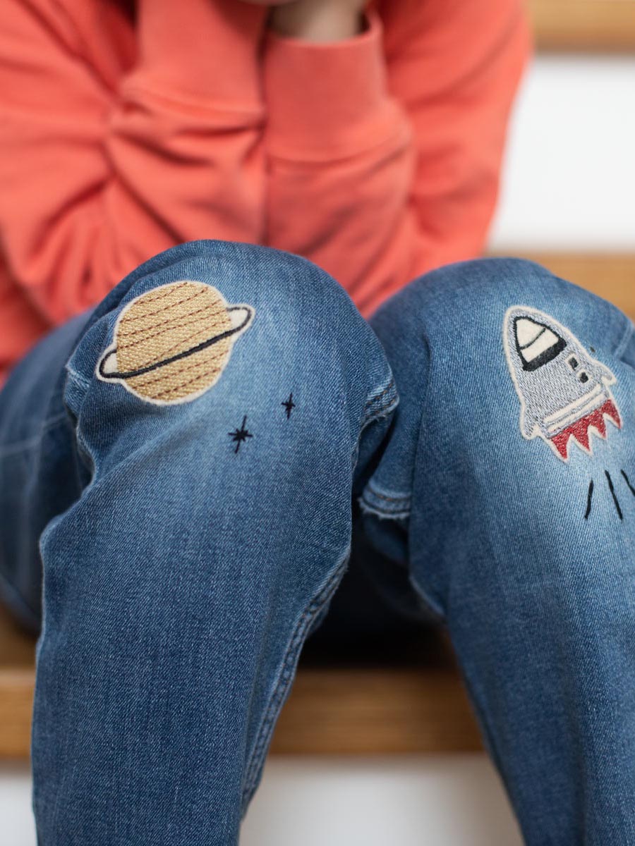 Kind sitzt auf der Treppe und hat zwei geflickte Knie von einer Jeanshose, rechts ist eine Rakete in grau und links ein Aufnäher Planet in gold