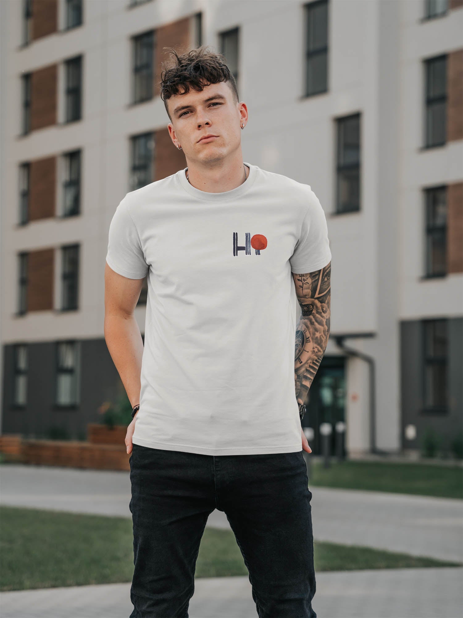 Mann mit einem weißen T-shirt auf dem ein Bügelbild mit dem Schriftzug hi gebügelt ist, er hat tattoos am arm und eine schwarze jeans n