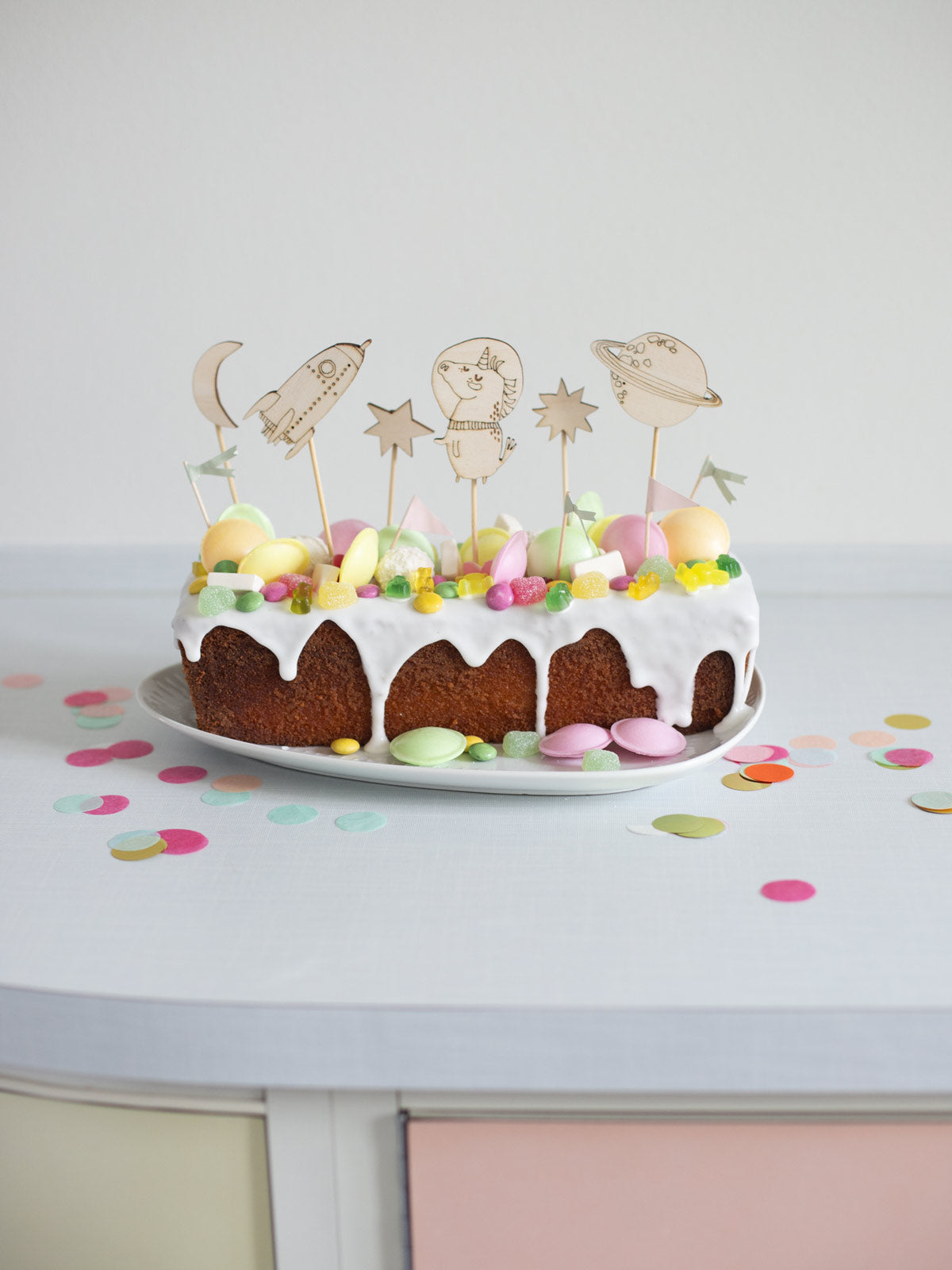 Zitronenkuchen verziert mit Glasur, Süssigkeiten und Cake Toppern aus Holz. Sie zeigen ein Einhorn im Weltall, eine Rakete, Planeten Mond und Sterne