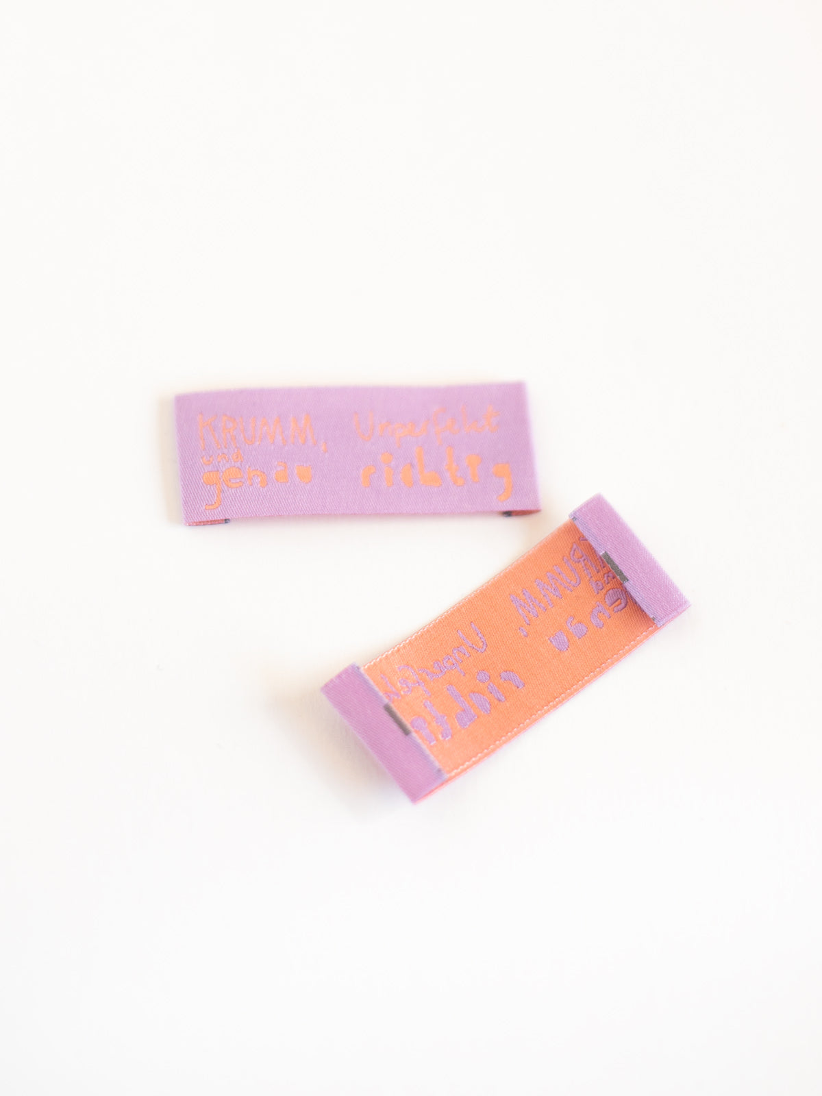 fliederfarbenes Nähetikett mit der Aufschrift "krumm, unperfekt und genau richtig" von Halfbird von vorne und hinten