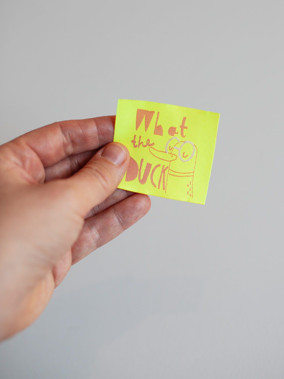 neongelbes Nähetiketten mit der aprikotfarbenen Aufschrift "what the duck" und Enten-Illustration von Halfbird