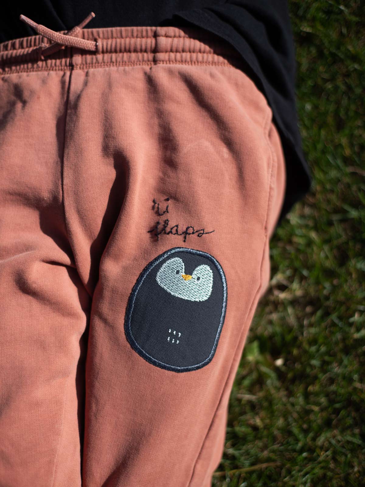 Kind liegt auf einer Wiese und trägt eine rotbraune hose auf der ein Pinguin Patch aufgebügelt ist und darüber steht gestickt hi flaps
