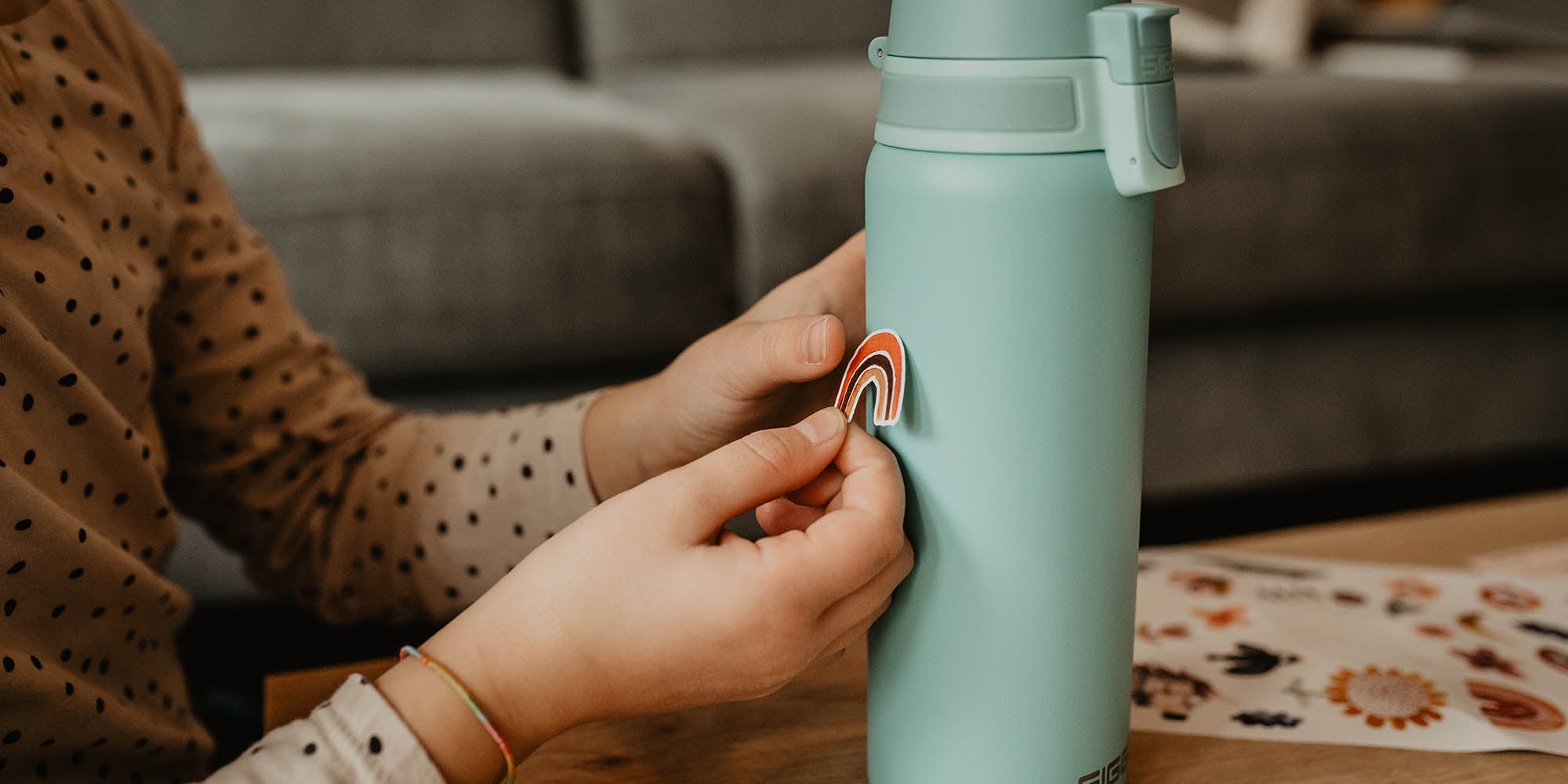 Kind klebt einen Regenbogen spülmaschinenfesten Aufkleber in rot, braun, rose auf eine Siggy Trinkflasche auf