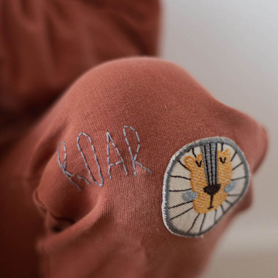 knie mit einer sweat hose und darauf ist ein bügelflicken löwe gebügelt der ein loch verdeckt, daneben steht roar gestickt