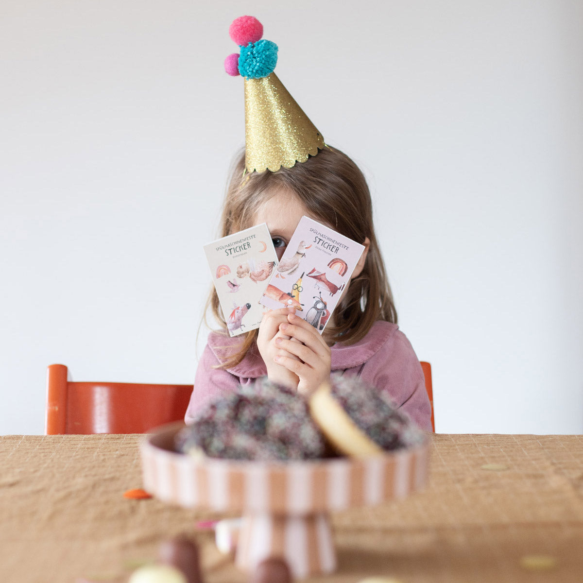 kind sitz vor dem tisch mit partyhut auf und hält zwei spülmaschinenfeste pocket bögen vor sein gesicht