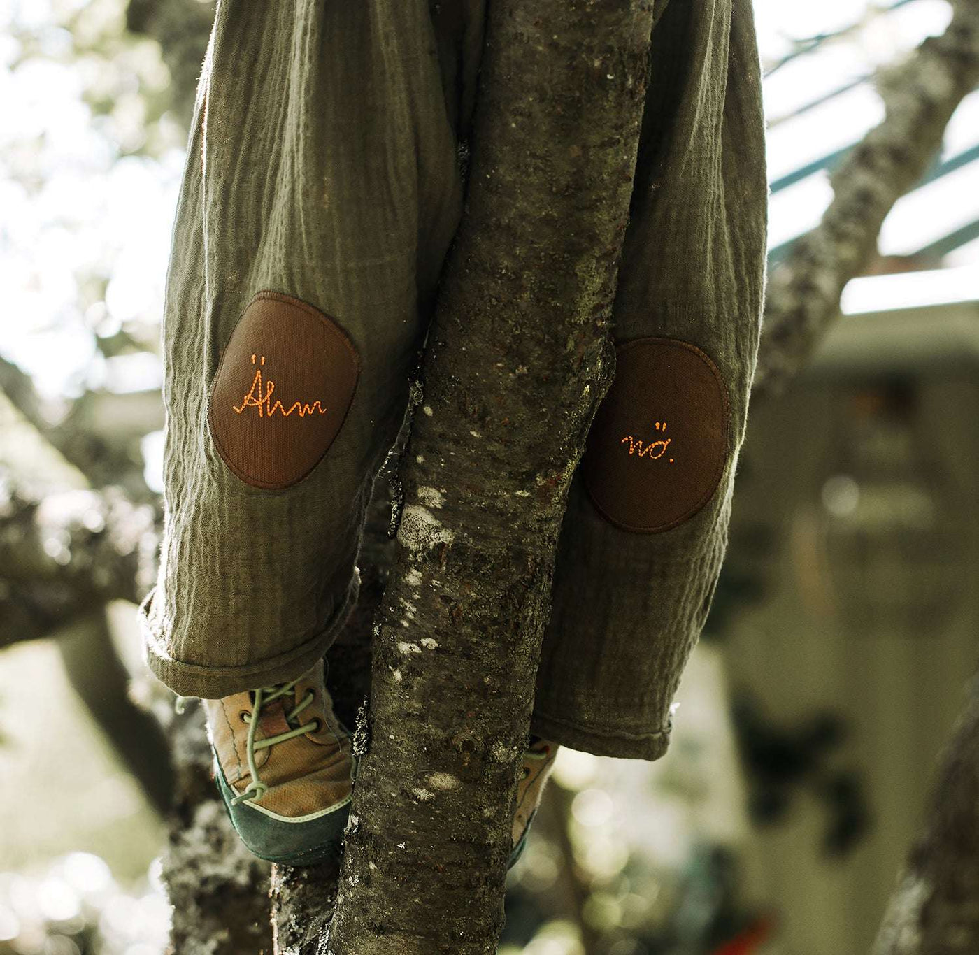 Kind klettert auf einem Baum udn hat eine musselin hose an mit zwei knieflicken in braun von halfbird mit dem schriftzug ähm nö