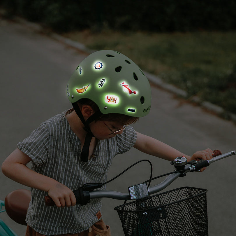 Kind steht neben seinem rad, es dämmert. es trägt einen grünen helm mit reflex stickern darauf die leuchten