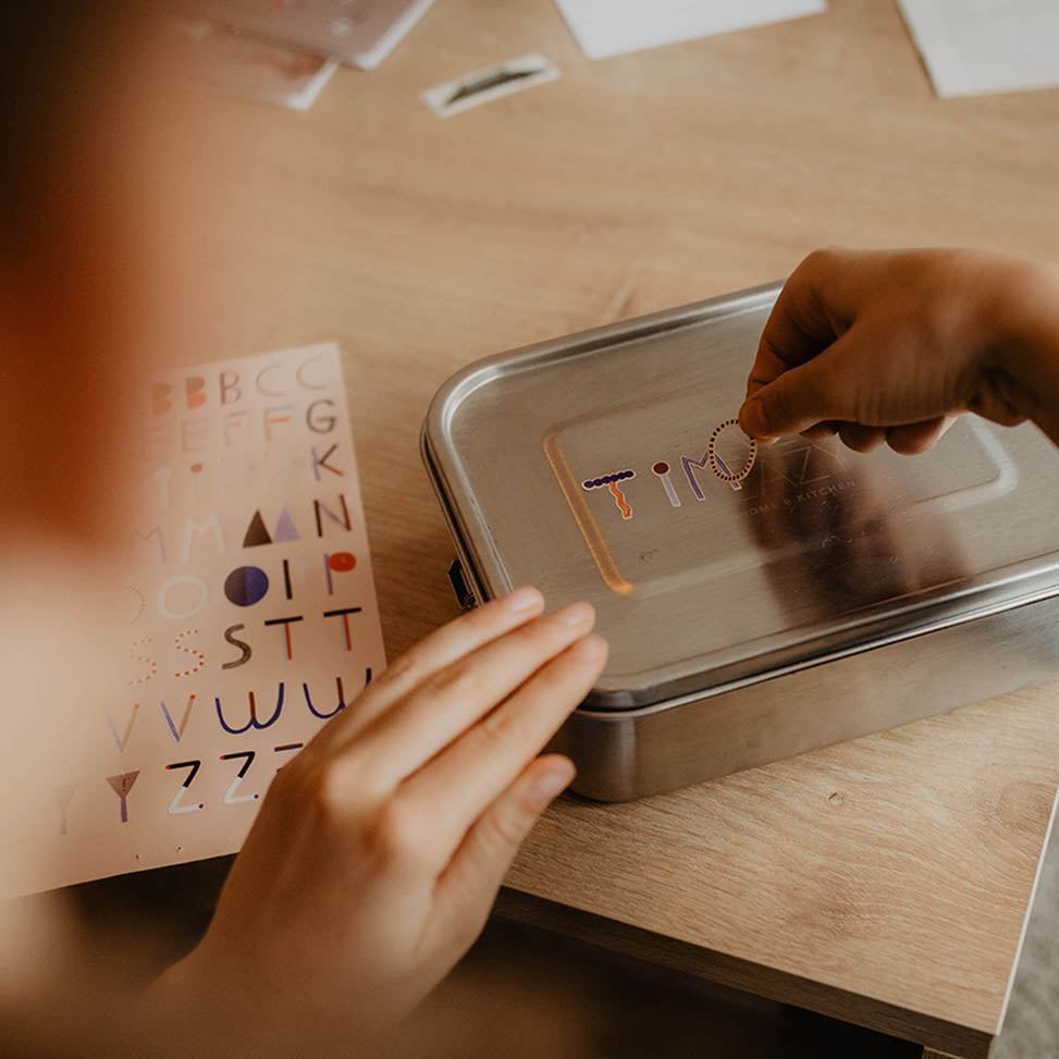 Kind klebt Timo mit Namensaufkleber auf eine Edelstahl Brotdose