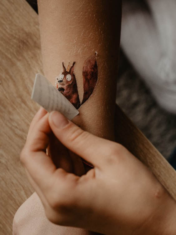 Kind zieht ein Trägerpapier vom Arm ab, darunter ist ein Kindertattoo Eichhörnchen von halfbird