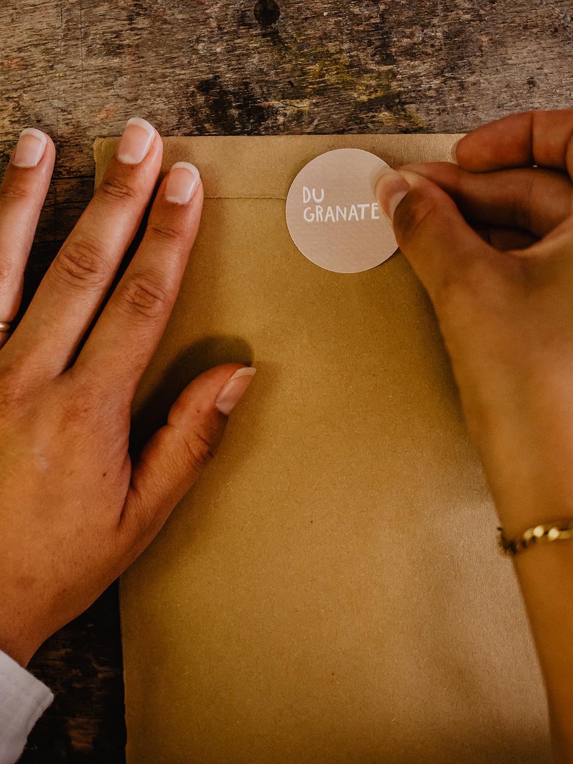 Hände kleben einen runden Sticker in zart rose mit dem Text du granate auf einen Umschag