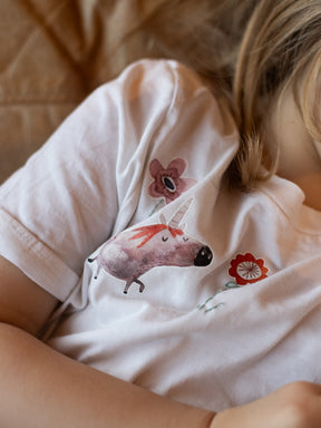 Kind liegt auf der couch und trägt ein weißes baumwollshirt mit einem rosa einhorn und blumen darauf, dazu wurden stiele gestickt