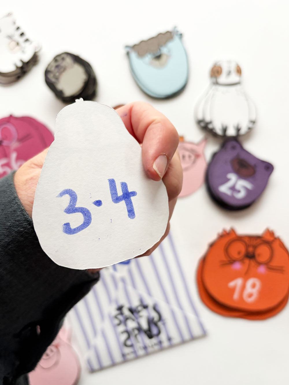 Hand hält Pinguin Spielkarte mit der Aufgabe 3 mal 4 auf der Rückseite aus dem Spielkartendeck von Halfbird. Im Hintergrund sichtbar, der gestreifte Umschlag und andere Kartenstapel, wie Schwein, Katze, Bär, Lama, Taube, Katze,...