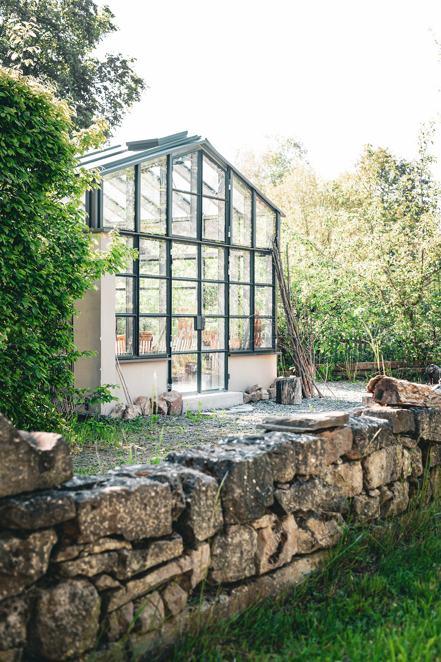 ein Gewächshaus steht in einem Naturgarten davor ist eine Bruchsteinmauer