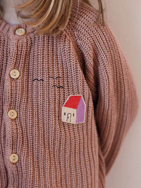 Kind trägt eine rose farbene STrickjacke mit einem Haus Aufnäher auf der Brust, der ein Loch verdeckt, daneben sind Vögel aufgestickt